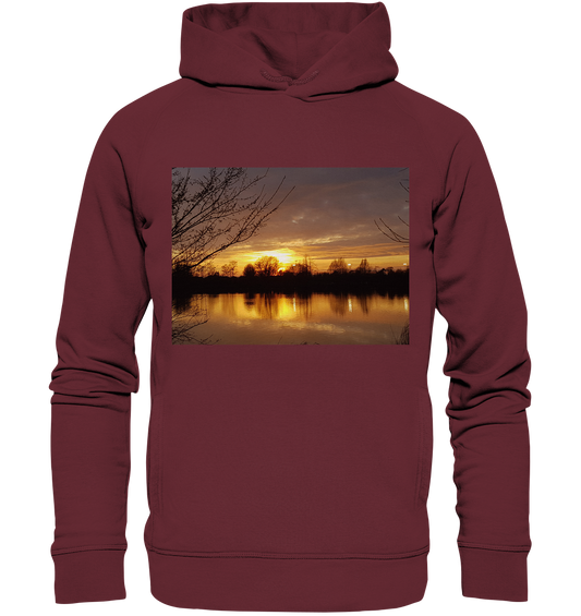 Der Abendspaziergang - Organic Fashion Hoodie von Silbers Magic Shop ist ein kastanienbrauner Hoodie, der einen ruhigen Sonnenuntergang über einem See zeigt, mit Bäumen, die sich vor einem farbenfrohen Himmel abzeichnen, der sich im Wasser spiegelt. Hergestellt in Bio-Qualität für diejenigen, die nachhaltigen Stil und Komfort schätzen.