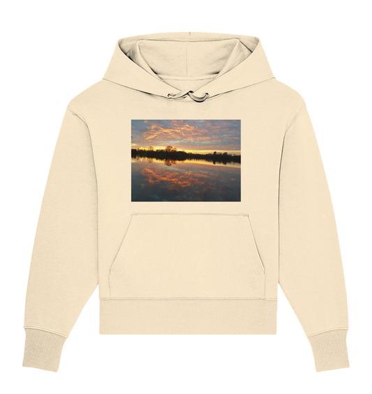 Der See am Abend - Organic Oversize Hoodie von Silbers Magic Shop ist ein beigefarbener, übergroßer Hoodie mit einer Vordertasche und einer Kapuze mit Kordelzug. Auf der Brust ist ein Bild zu sehen, das einen ruhigen Sonnenuntergang über dem Wasser zeigt, komplett mit einer Baumsilhouette und leuchtenden Reflexen in Orange- und Rosatönen. Hergestellt aus Bio-Baumwolle für zusätzliche Nachhaltigkeit.